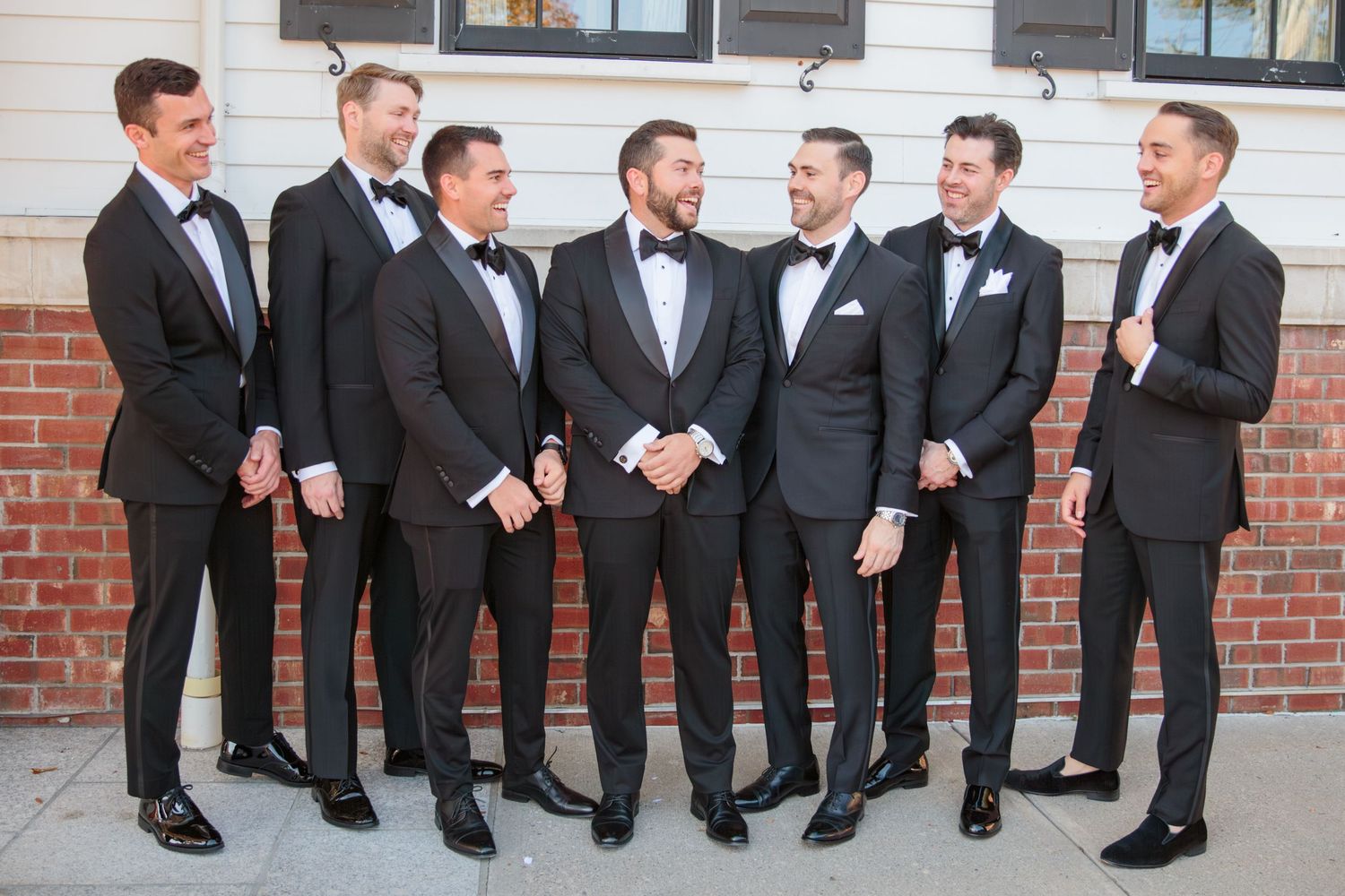 A group of groomsmen in black tuxedos and bow ties gather for a casual moment outside a brick building.