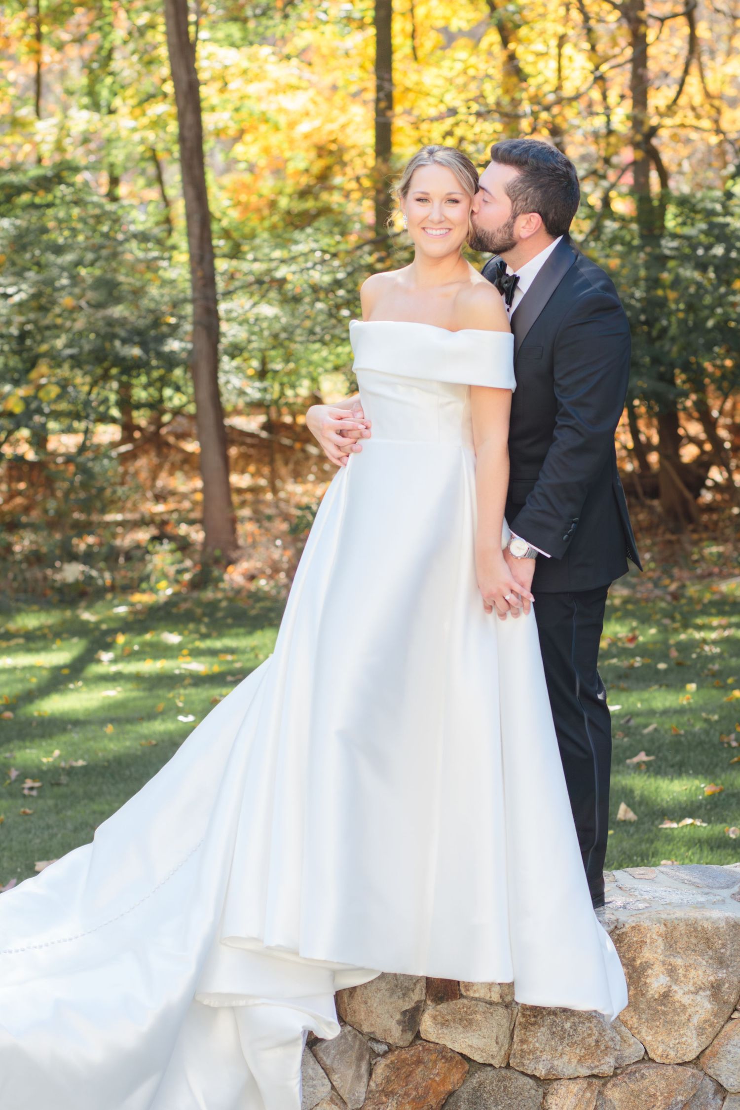 A wedding couple shares romantic moments on a stone wall surrounded by autumn foliage in a garden setting.