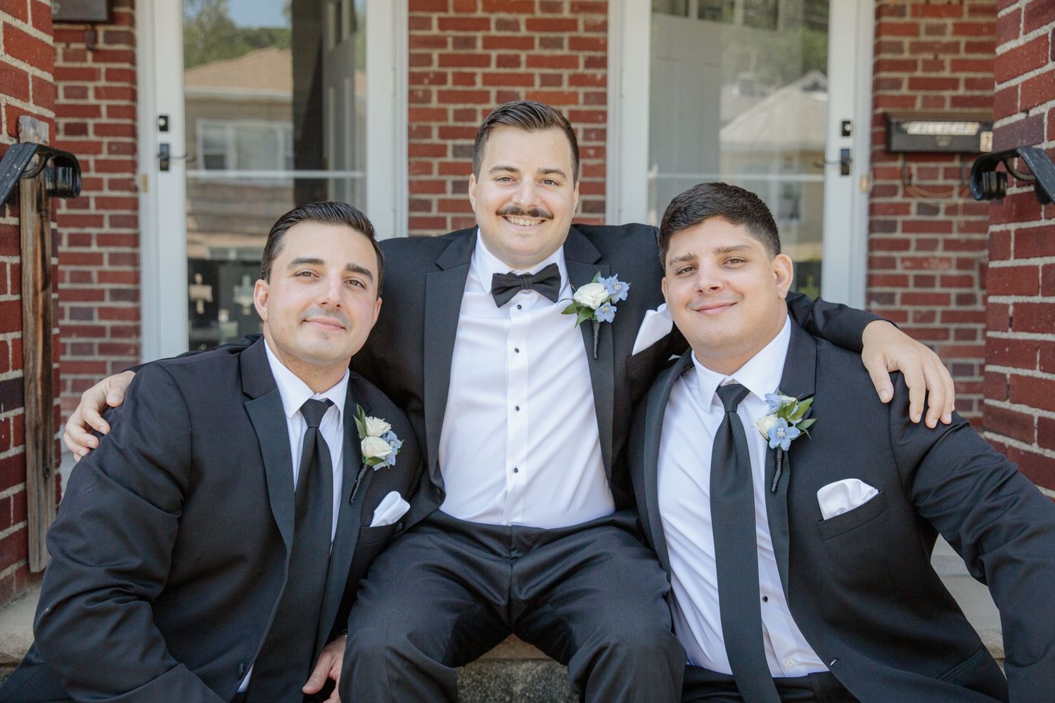 Groomsmen and Groom in black suits and ties pose together in front of a brick building.