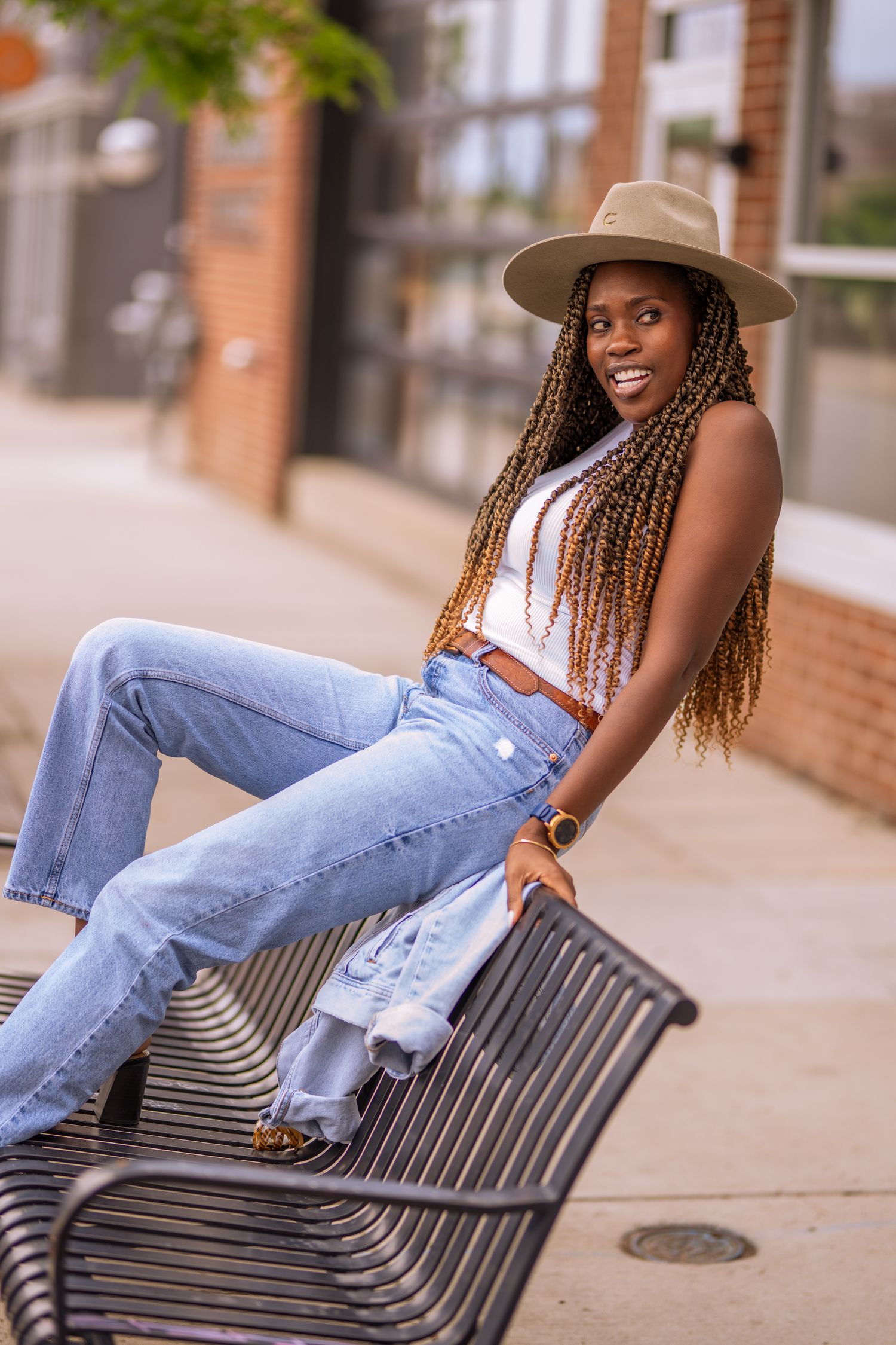 A stylish person in a beige hat and white top lounges casually on a metal bench wearing ripped blue jeans.
