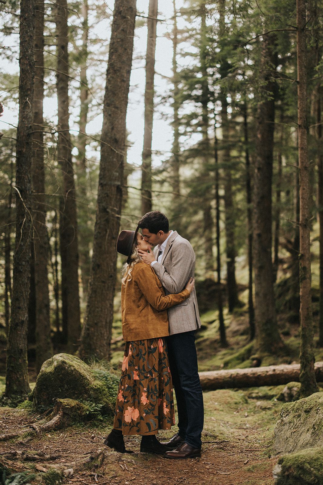 A couple shares an intimate moment on a forest path surrounded by tall pine trees and mossy rocks.