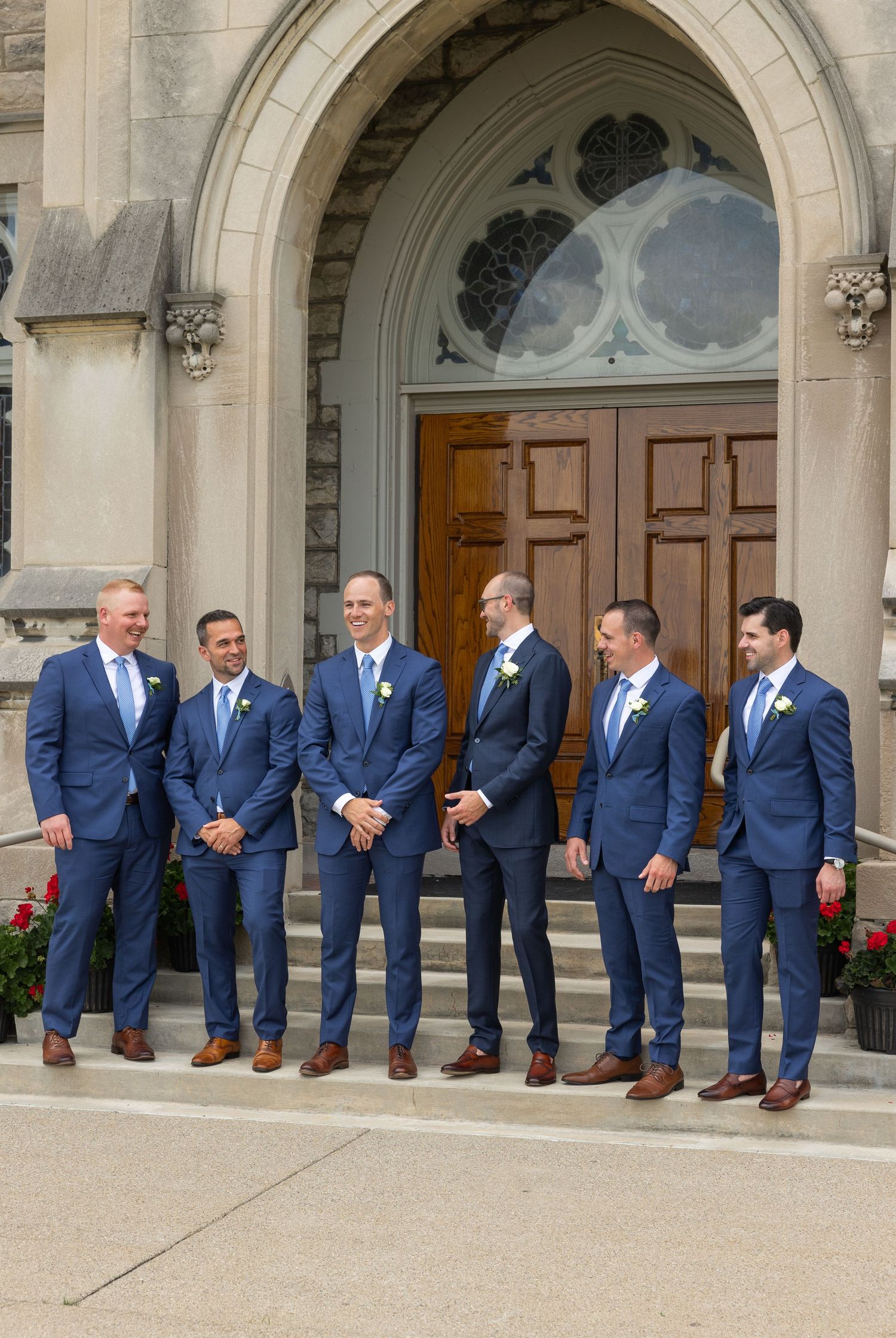 Groomsmen in matching navy blue suits and brown shoes stand together on church steps beneath an arched doorway. Rachel Martinez Photography