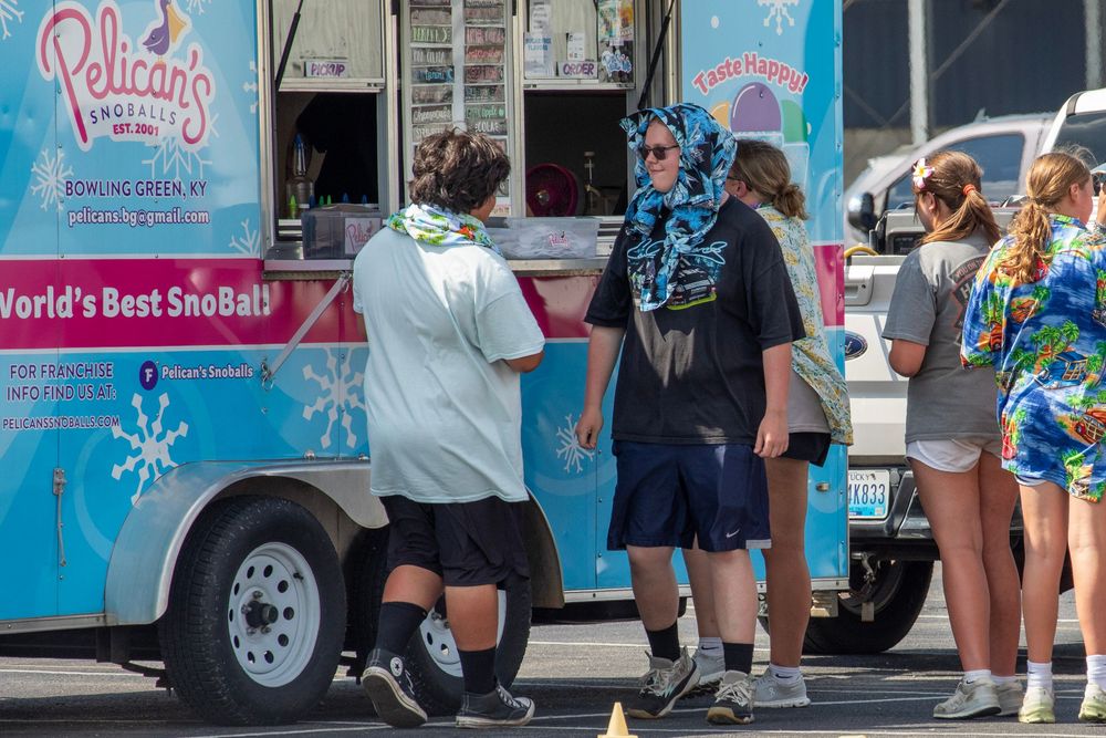 A line of customers wait to order from a light blue Pelican's Snowballs food truck on a sunny day.