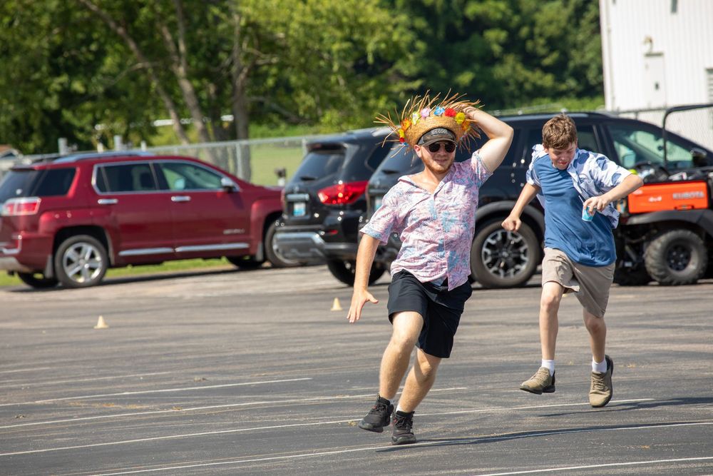 Two students playfully chase each other across a parking lot on a sunny day with parked cars in the background.