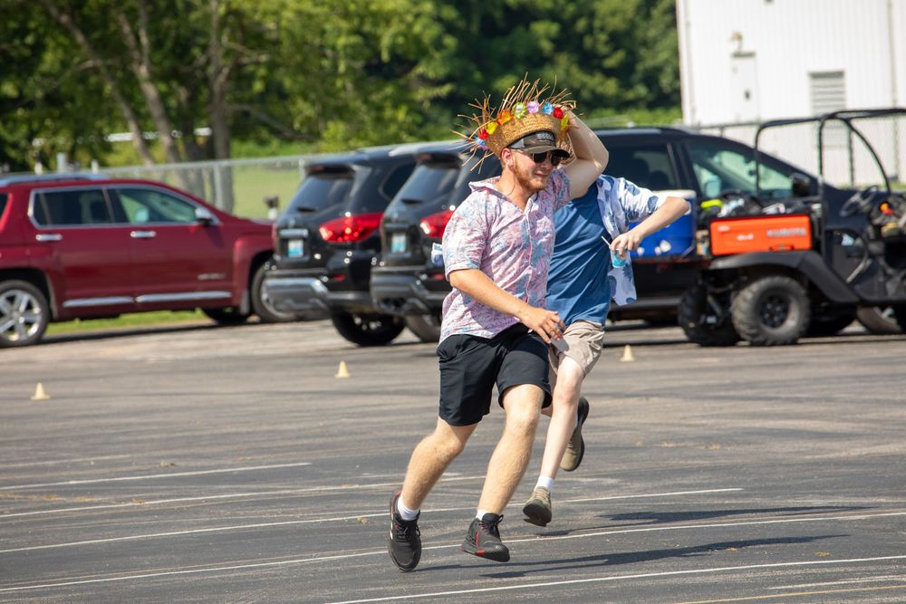 Action shot of a runner leaping energetically across a sunny parking lot with parked cars in the background.