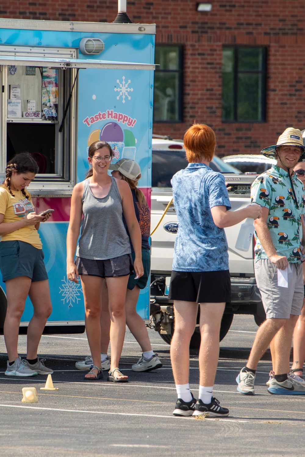People waiting in line at an ice cream truck parked in front of a brick building on a sunny day.