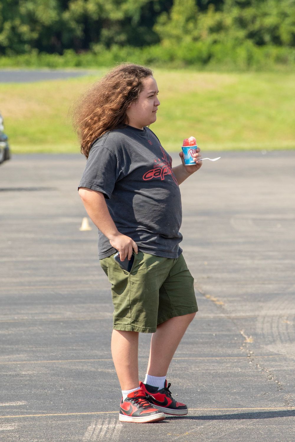 Someone in a dark t-shirt and green shorts walks across an outdoor parking lot in summer.