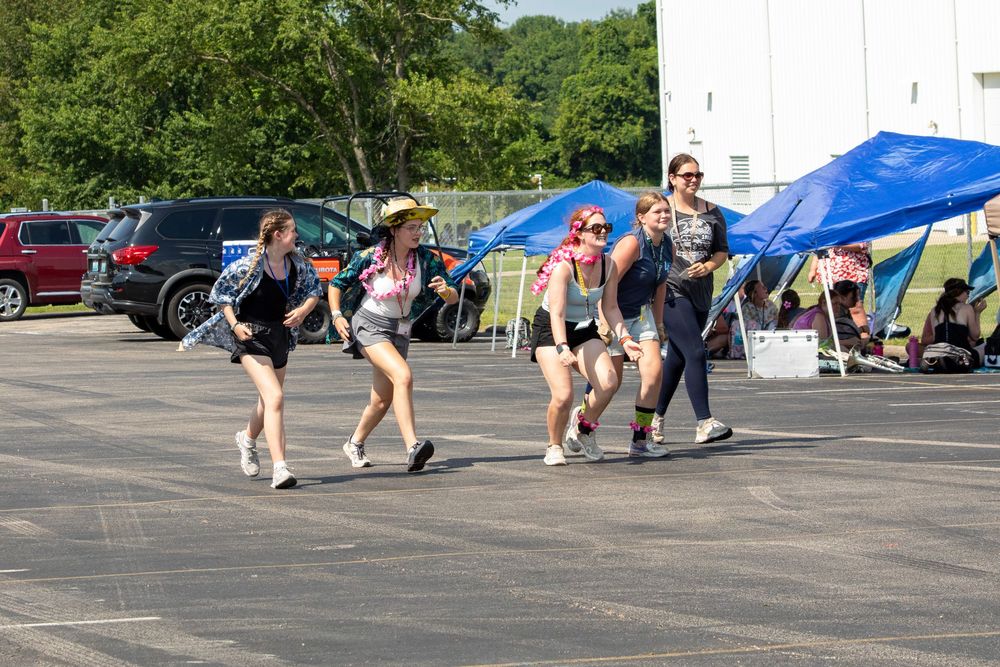 Athletes run along a paved asphalt path near green trees and blue tents during an outdoor race event.