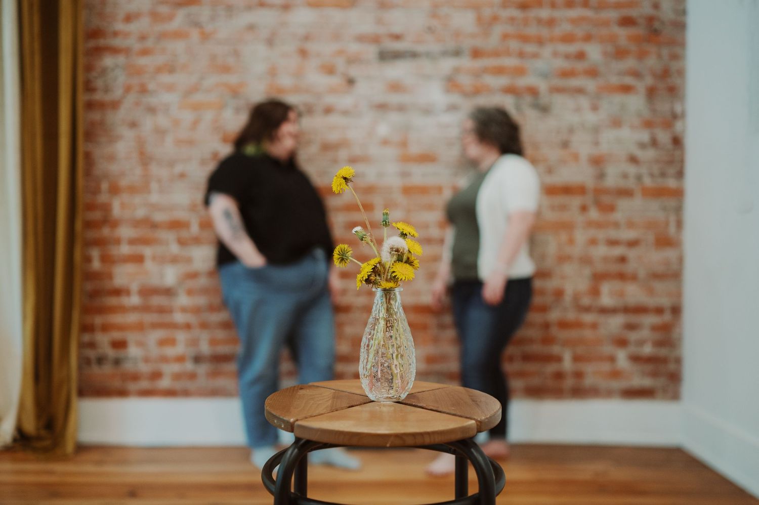 Vase of sunflowers on wooden table with brick wall background and figures standing in soft focus.