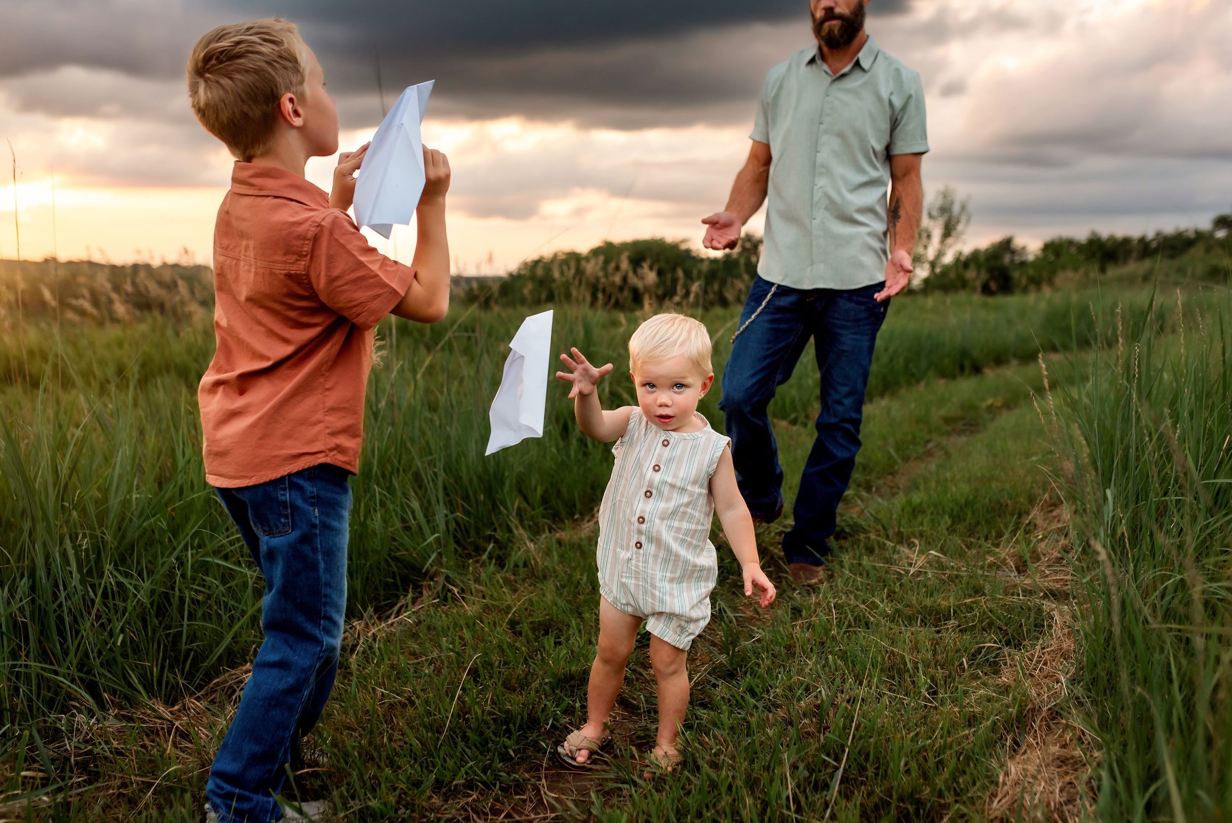 A family walking through a tall grass field at sunset while holding paper airplanes.