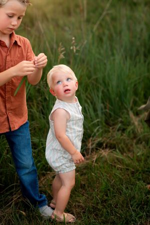 Two figures stand together in tall grass during golden hour, one wearing a brown shirt and jeans, the other in striped romper.