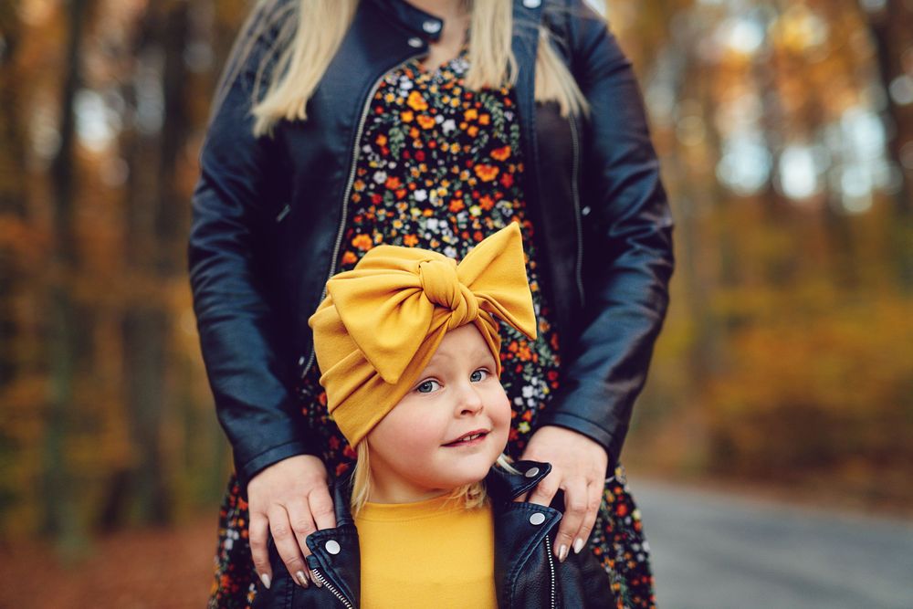 A person in a navy coat holds a child wearing a yellow bow in an autumn forest setting.