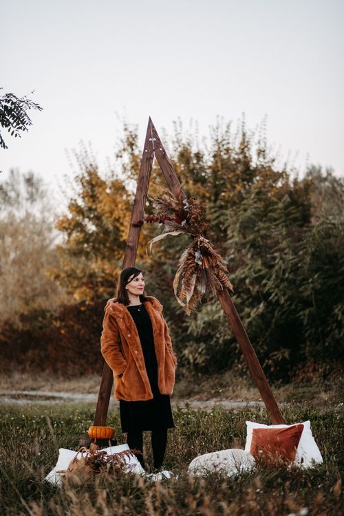 Rustic autumn outdoor scene with brown corduroy jacket, pumpkin, and decorative wooden cross against natural foliage.