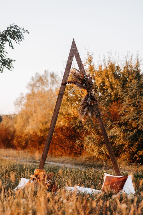 Rustic wooden A-frame structure in autumn field with white cushions and golden foliage in background.