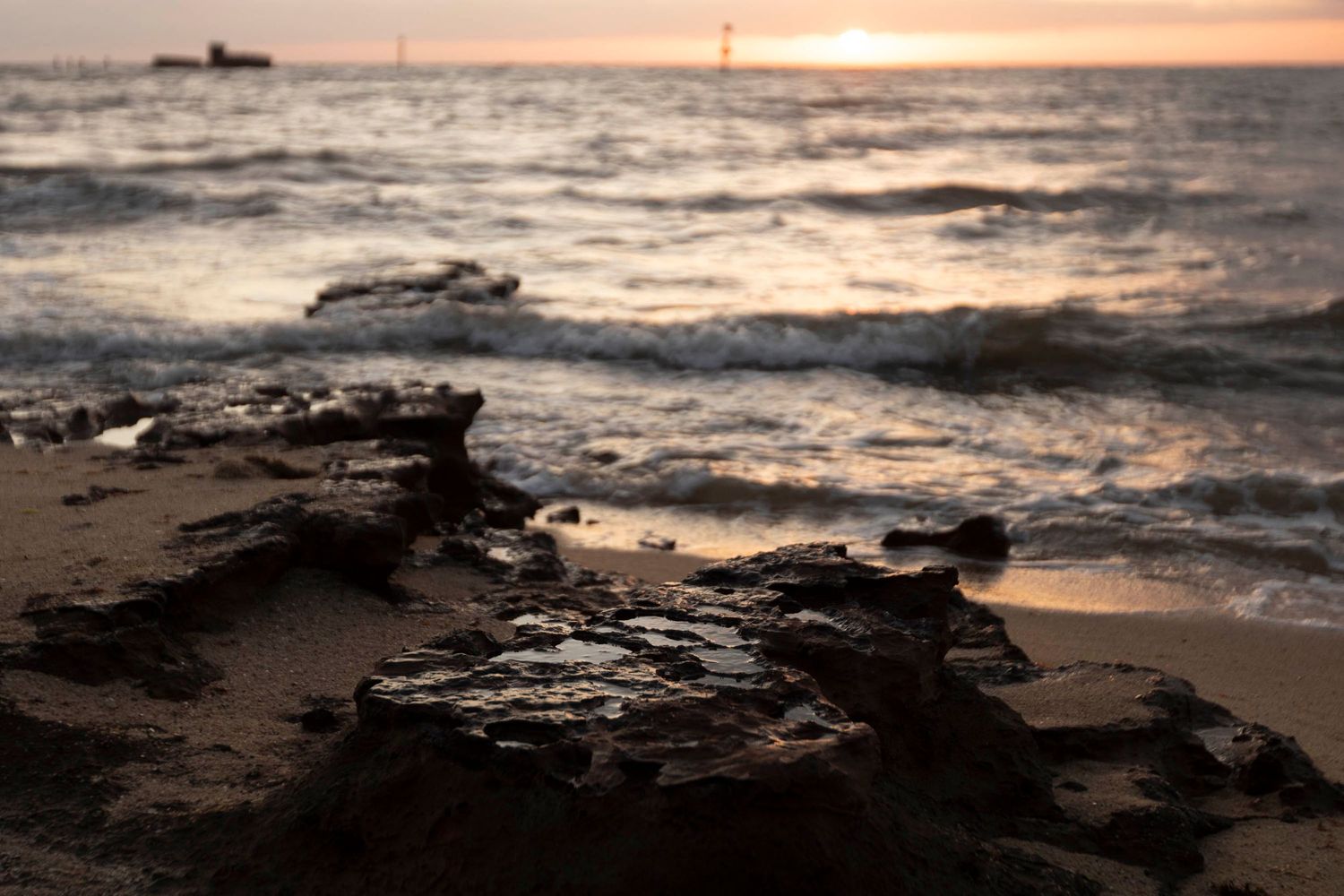 Rocky beach shoreline at sunset with waves crashing against dark stones in golden light.