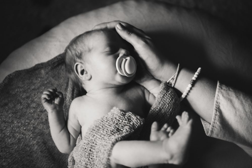 Black and white image of a crying newborn baby wrapped in knitted fabric being comforted by gentle hands.