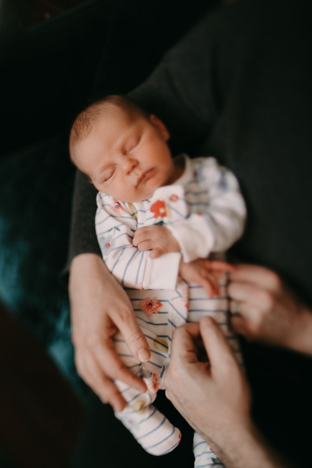 A sleeping infant wearing a striped onesie being gently cradled by adult hands against a dark background.