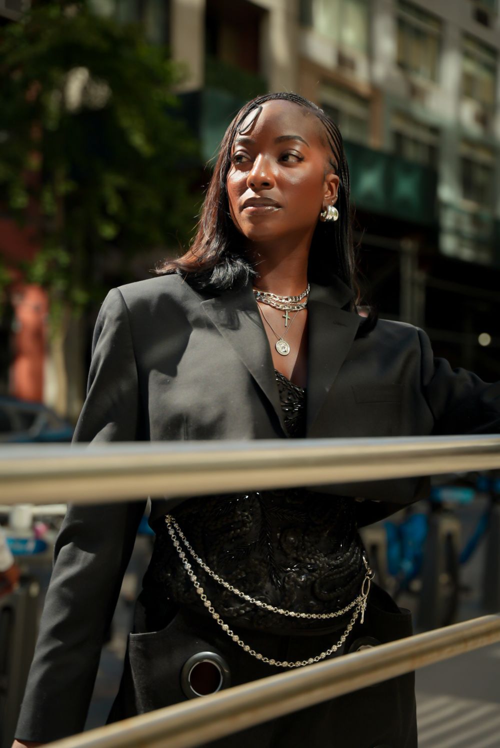 A person in a black blazer and jewelry poses confidently against a railing in an urban setting.