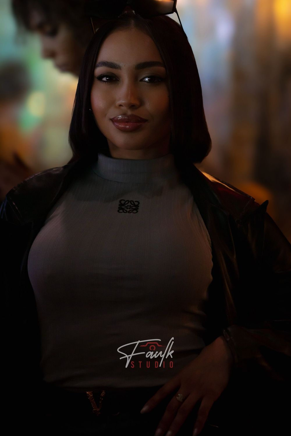 A person wearing a white top and black jacket poses in moody lighting with a soft smile.