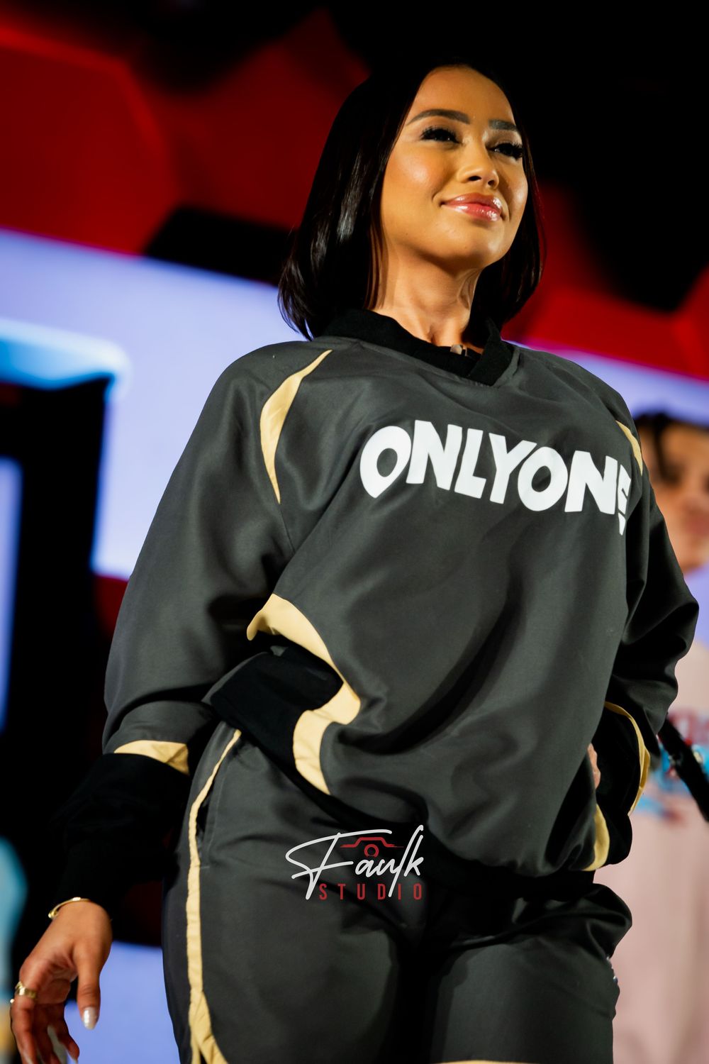 Person wearing a black sweatshirt with gold accents performs on stage under dramatic lighting.
