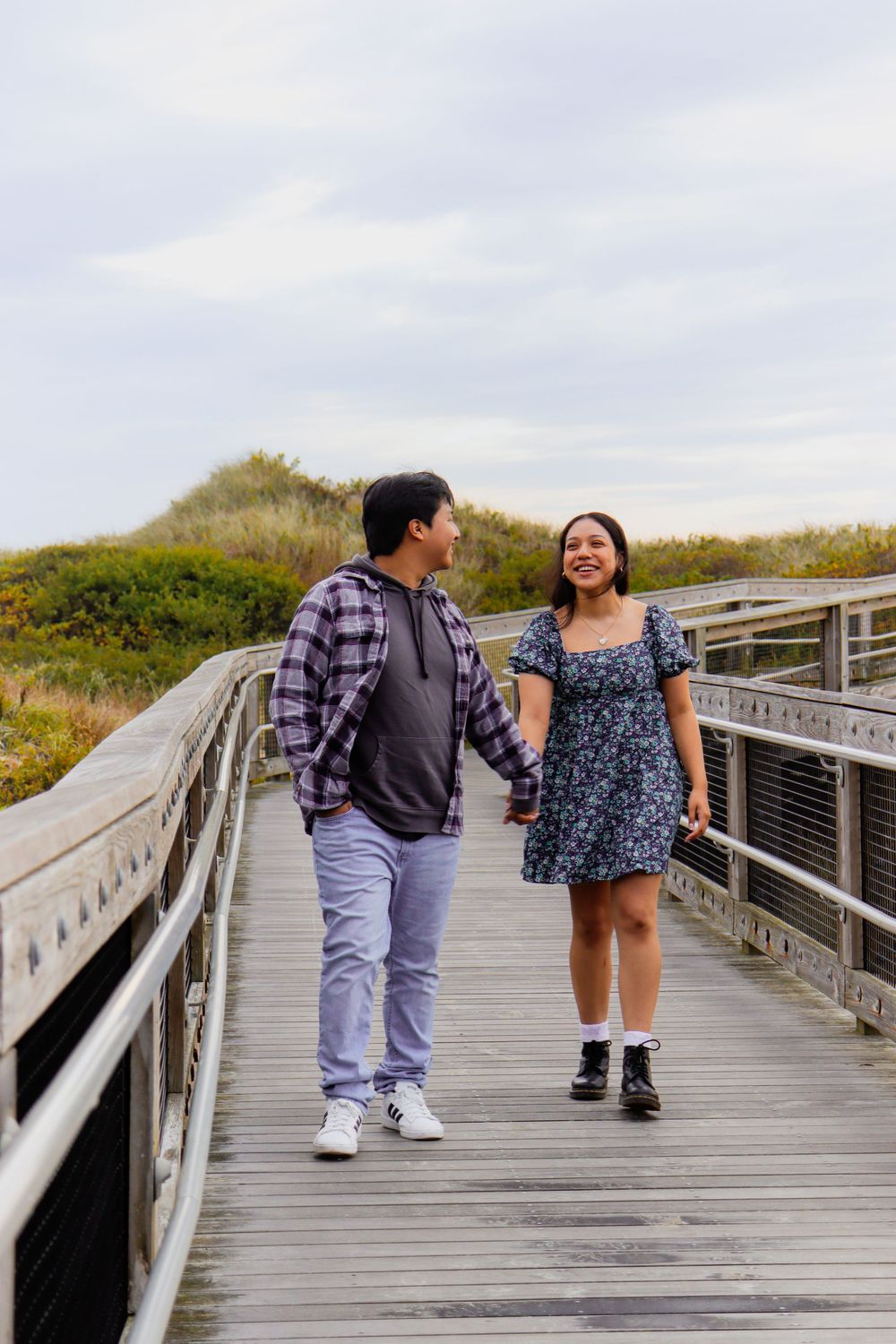 A couple walks together on a wooden boardwalk surrounded by coastal vegetation.