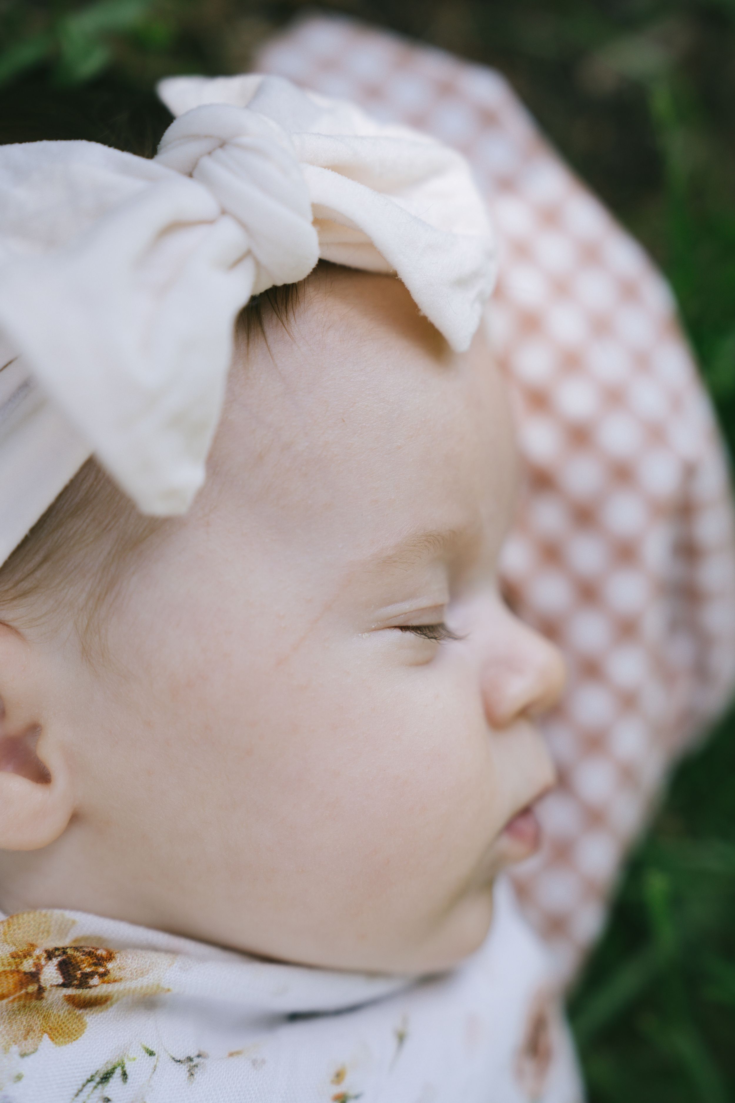 A peaceful moment captured of a baby wearing a white bow headband against greenery.