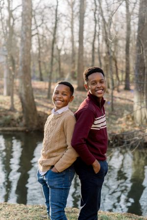 Two people posing back-to-back by water with bare trees in winter woodland setting.