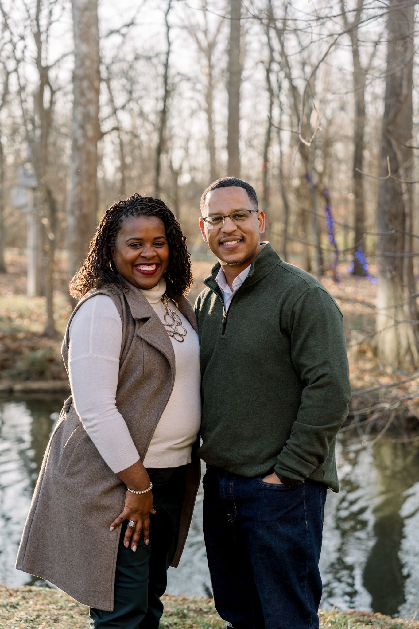 Couple portrait in casual winter attire standing by scenic pond in wooded area.