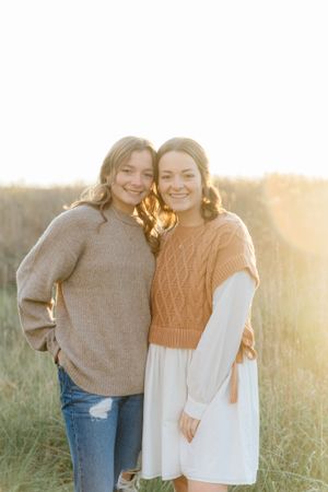 Two people in cozy sweaters embrace while standing in a sunlit field at golden hour.