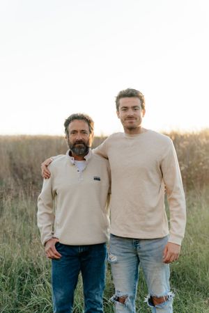 Two people in neutral sweaters pose together in a field at sunset.