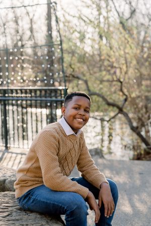 A young person in a tan cable knit sweater sits casually outdoors on a sunny day.
