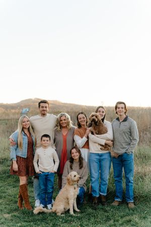 Extended family poses for outdoor portrait with golden retriever dog in grassy field at sunset.