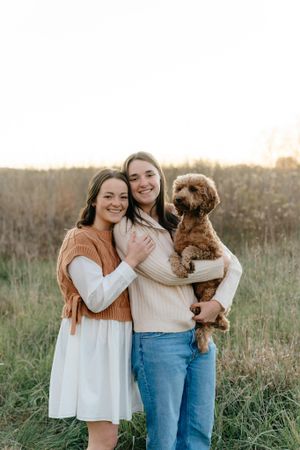 Two women embrace while holding small brown dog in grassy field during golden hour.