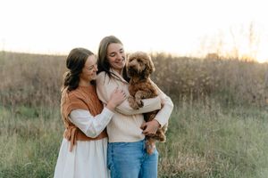 Two friends share joyful moment with curly-haired puppy in autumn field setting.