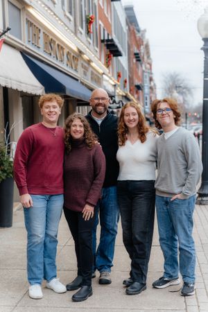 Group of five people in casual sweaters pose together on sidewalk in front of storefronts.