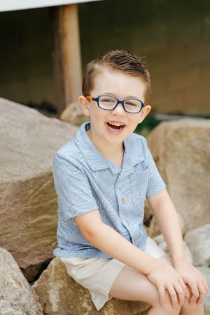 Young child in blue button-down shirt and glasses sitting on rocky surface outdoors.