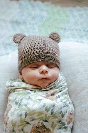 A sleeping infant wrapped in floral swaddle wearing a gray knitted bear hat with tiny ears.
