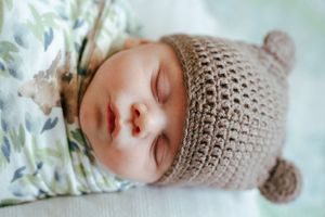 Close-up of a peaceful sleeping baby wearing a tan crocheted bear hat against blue background.