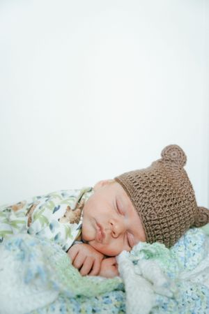Sleeping newborn in beige bear hat swaddled in floral blanket against white background.