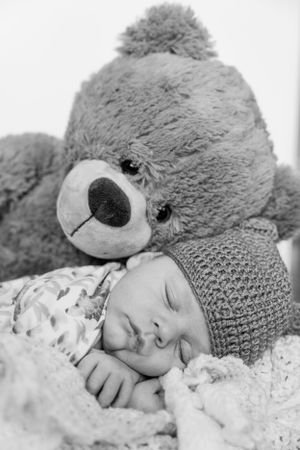 Black and white photo of sleeping baby with knit hat next to large teddy bear.