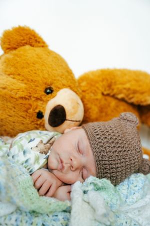 Sleeping baby in bear hat cuddled next to large golden teddy bear on light blue blanket.