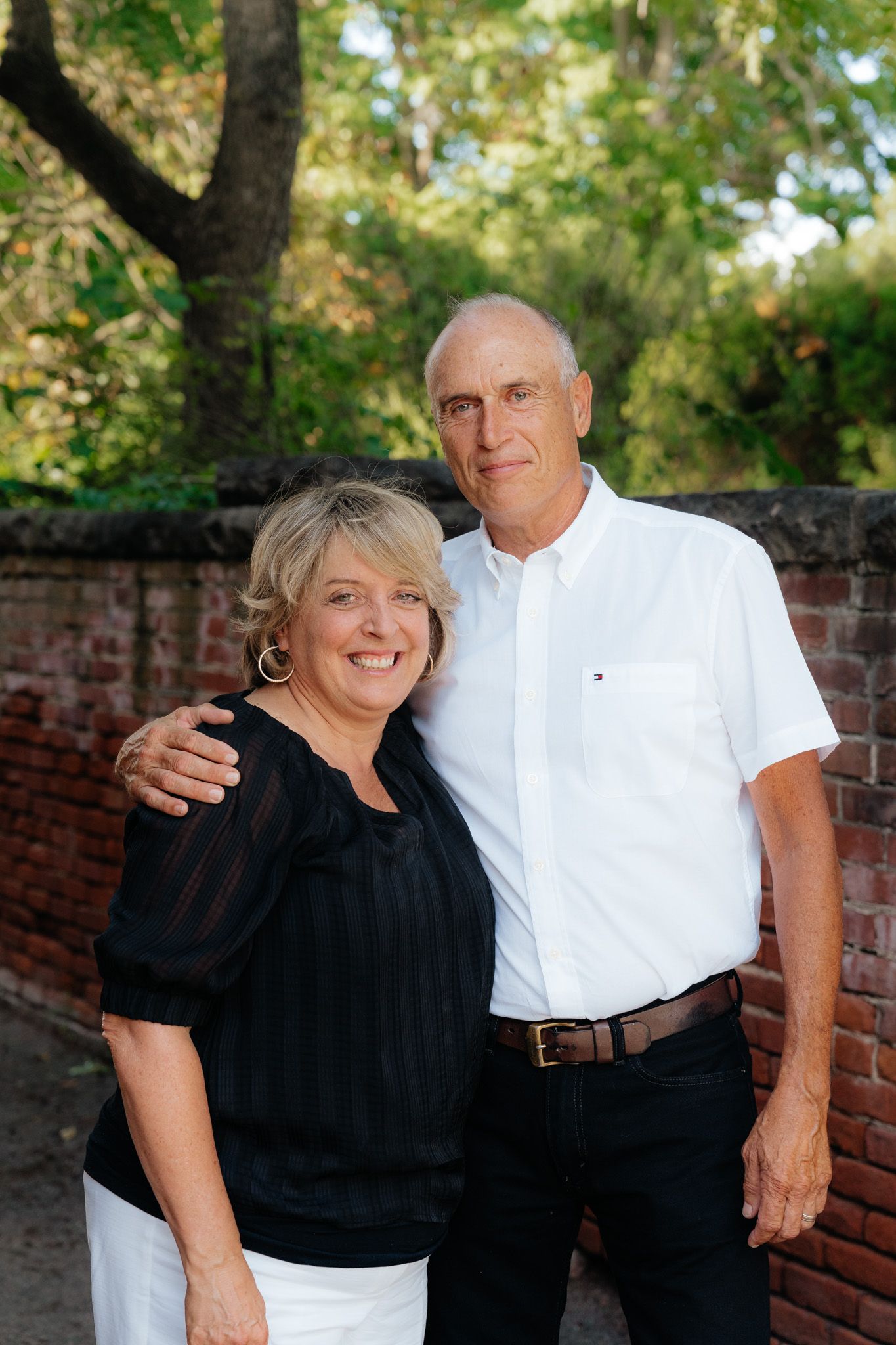 Couple in casual attire standing together against brick wall and green trees.