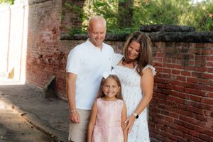 A family of three poses together against a rustic brick wall in coordinating white and pink outfits.