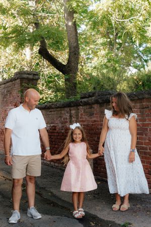 A family holds hands while standing in front of a brick wall and trees on a sunny day.