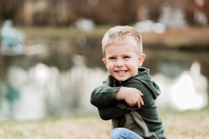 Young child in green hoodie kneeling outdoors by a pond.