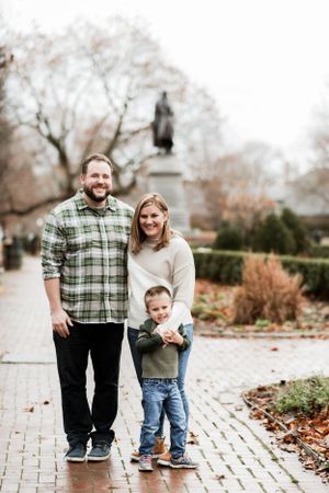 Family of three standing together on brick path in autumn park.