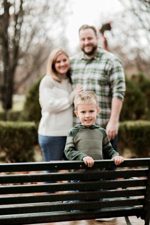 A young child leans on a wooden bench in a garden while a couple smiles in soft focus behind him.
