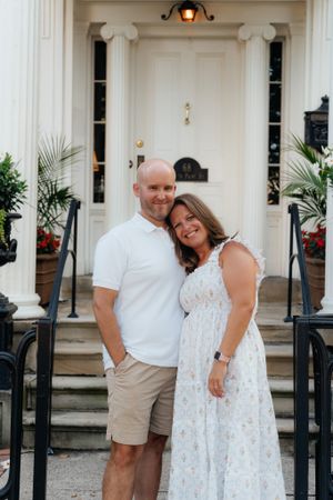 A couple poses together on steps in front of an elegant white doorway.