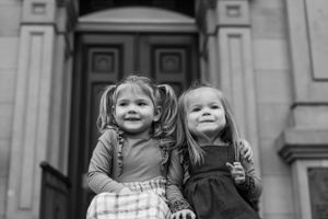 Two young children in dresses stand together smiling in front of a grand doorway.
