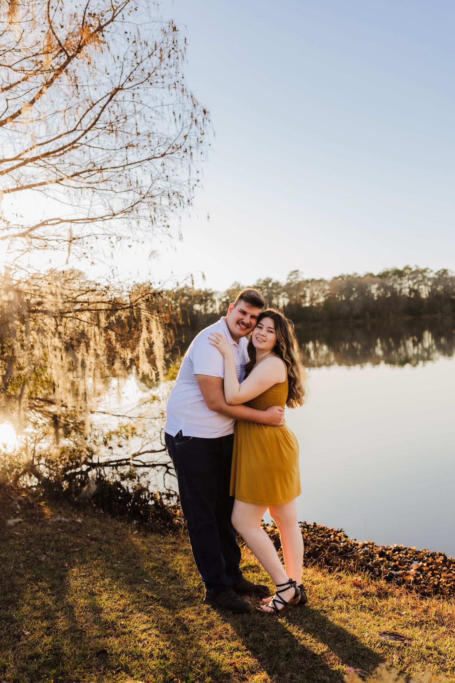 Romantic golden hour portrait by lakeside with warm sunlight filtering through autumn trees.